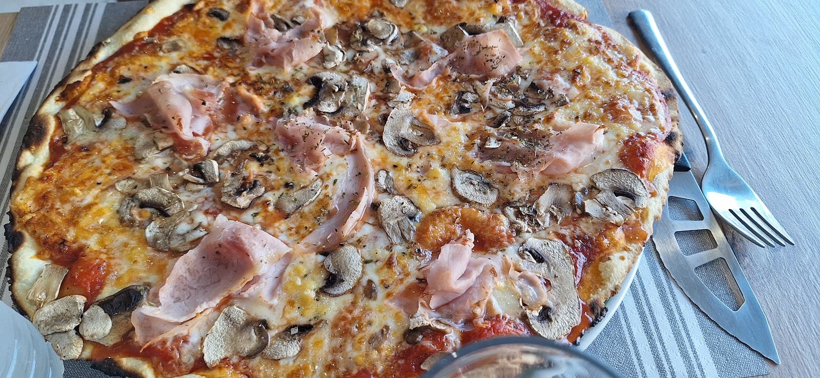 Pizza Reine au jambon et aux champignons, dorée au four à bois, posée sur un set de table rayé avec couteau et fourchette