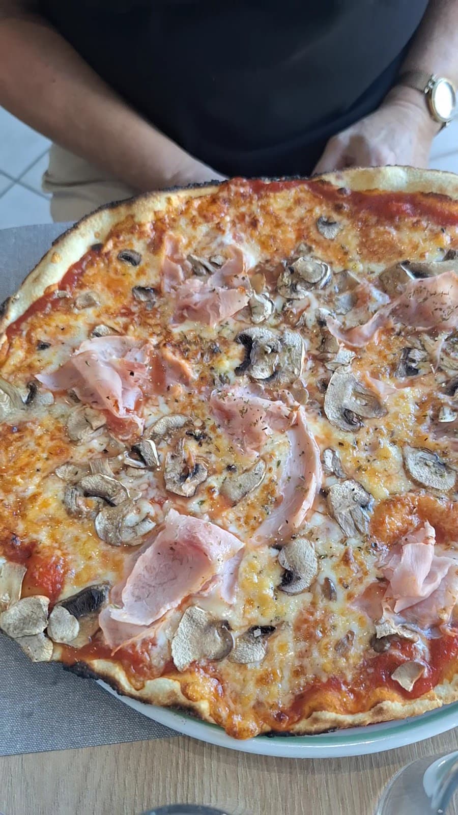 Une grande pizza Reine au jambon et aux champignons, tenue sur son assiette par un membre de l’équipe en tablier noir