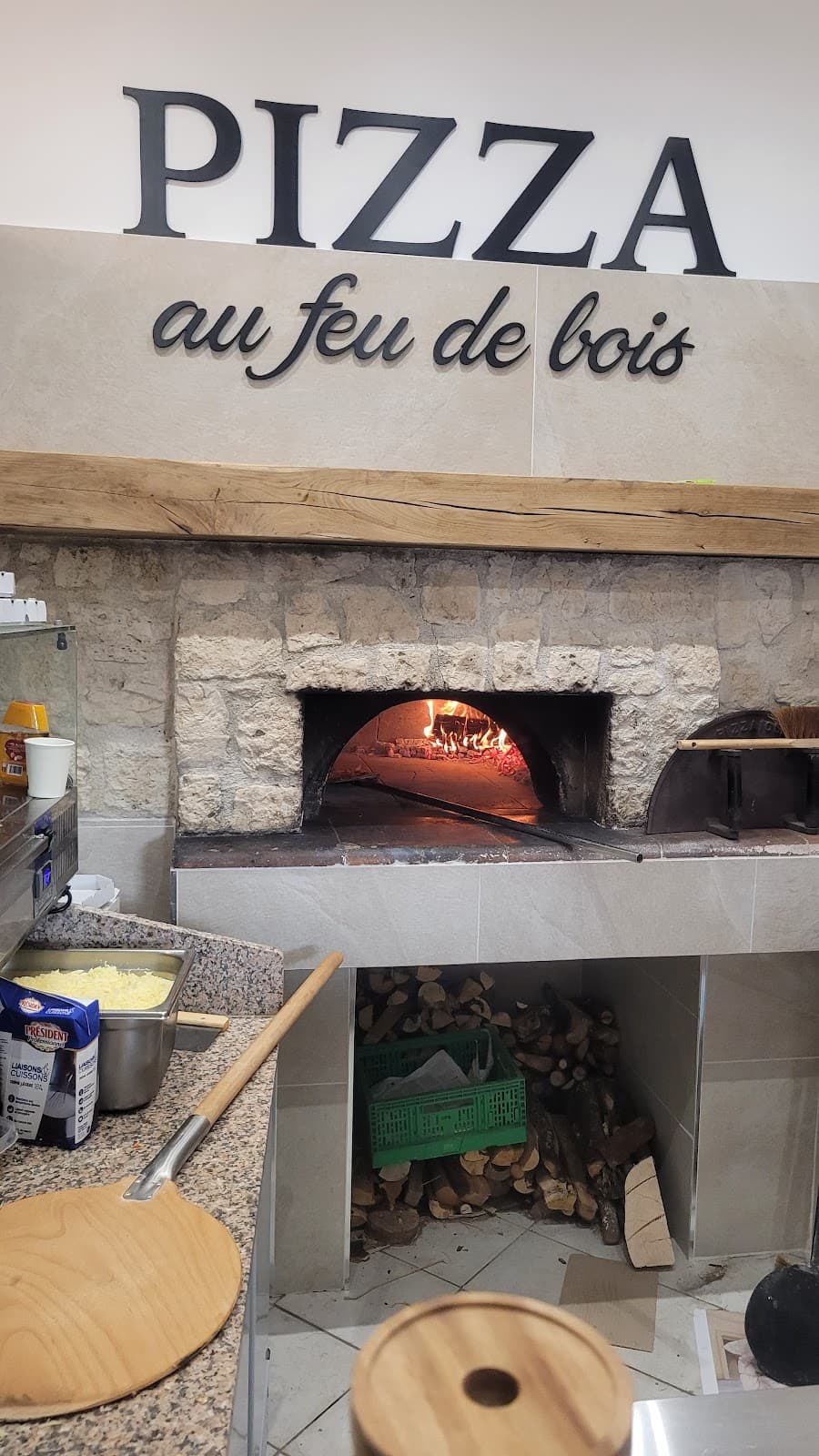 Le four à bois en pierre de La Casa di Stefano, flamme allumée dans la gueule du four et bûches empilées en dessous, sous l’enseigne « Pizza au feu de bois »